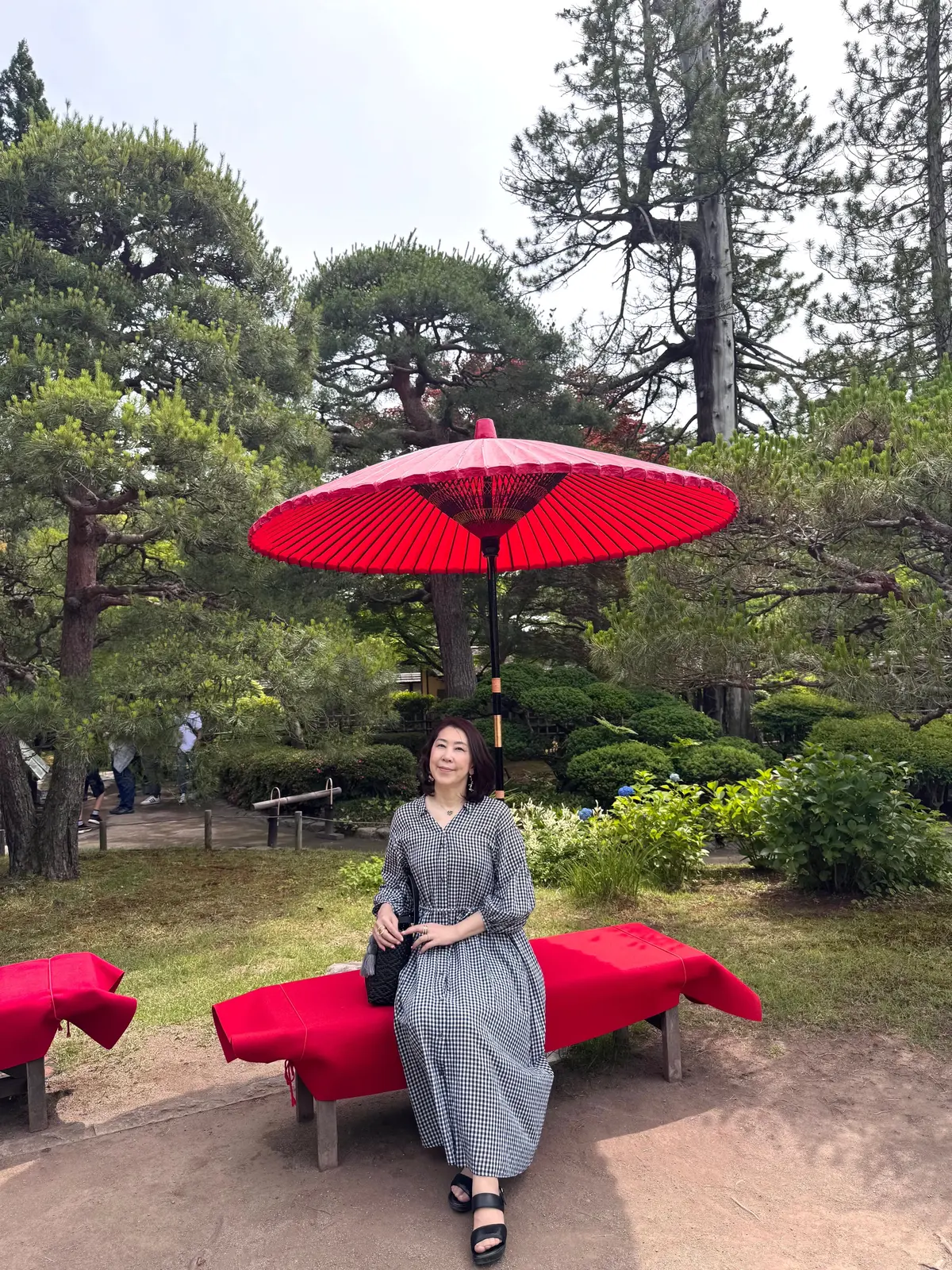赤い傘が立つ芝生の上に、チェック柄のワンピースを着た女性が赤いベンチに座っている。背景には松の木や低木が広がる庭園。