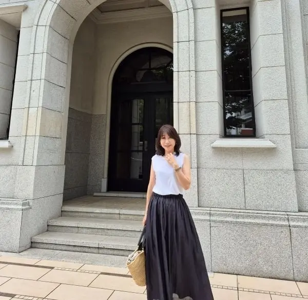 石造りの建物の前に立つ女性。白いタンクトップに黒いロングスカート、茶色のカバンを斜め掛けしている。髪は肩までの長さの茶色。建物の入り口のアーチが背景に見える。