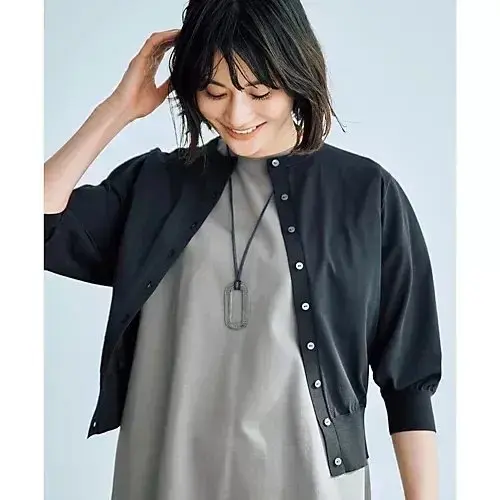 黒い襟なしのカーディガンを羽織った女性。グレーのワンピースを着ており、長めのネックレスを着用。右手を頭に当て、微笑んでいる。背景は淡いブルー。