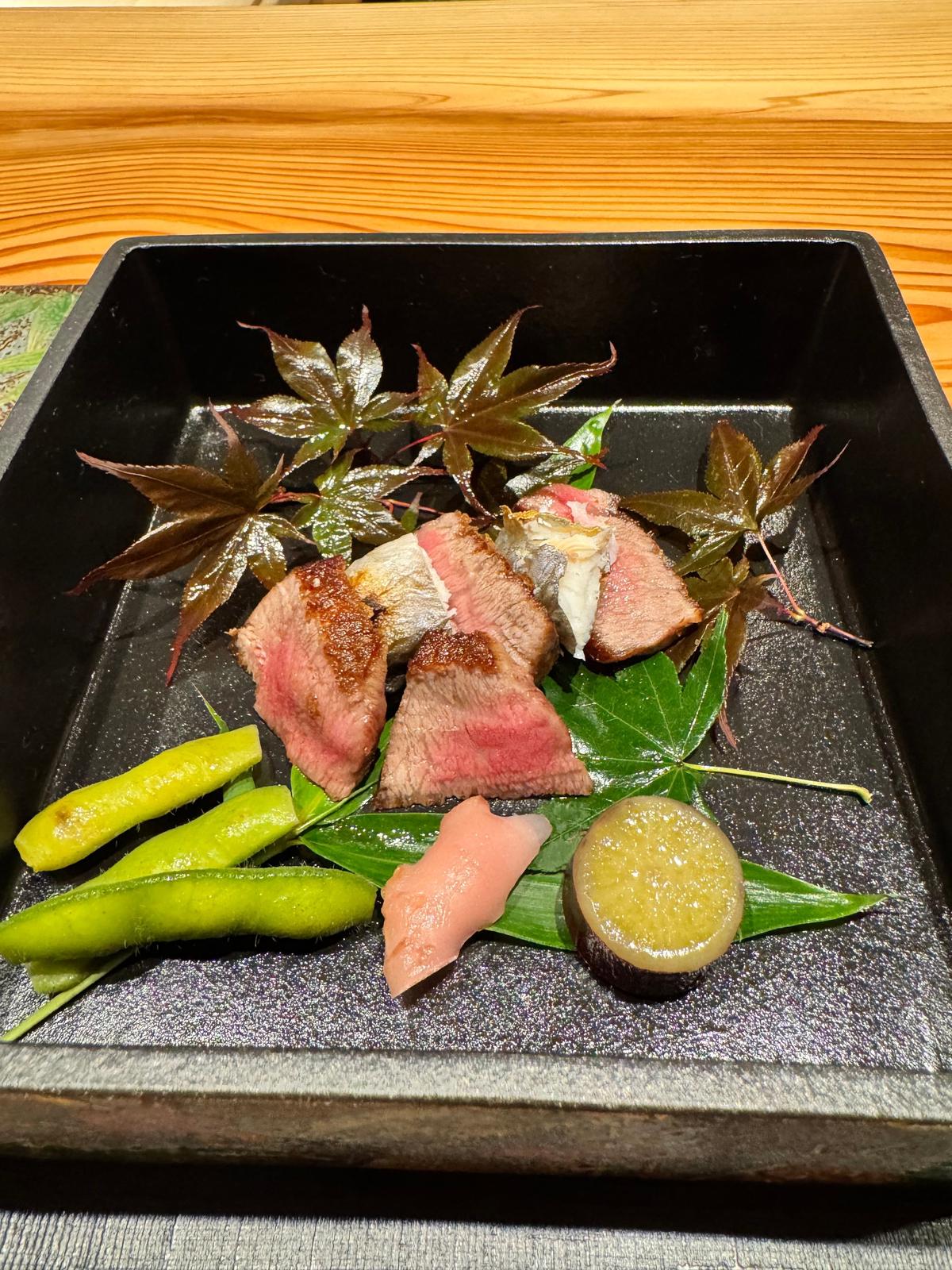 黒い正方形の皿に盛り付けられた、赤身の焼肉と、エディブルフラワー、スナップエンドウ、紫色の柑橘類。