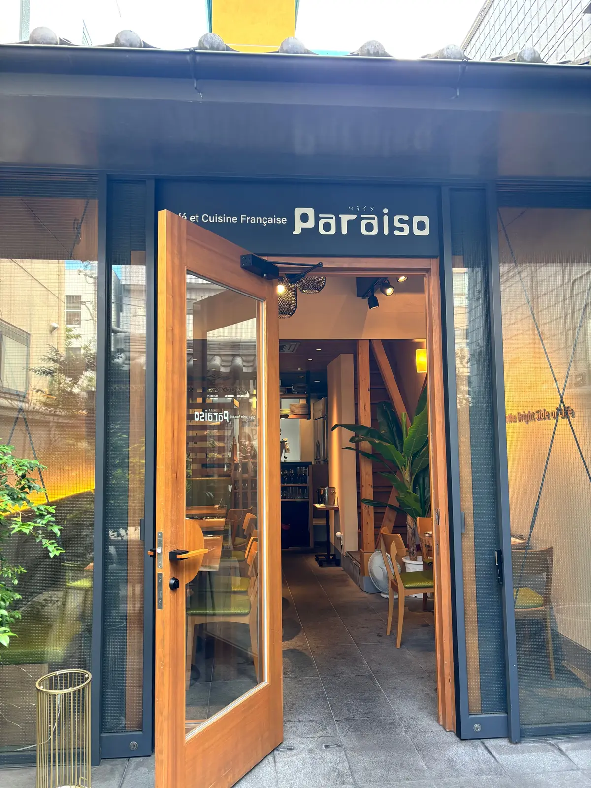 カフェ「パライソ」の入り口。木製のドアが開き、店内にはテーブル席と緑の植物が見える。