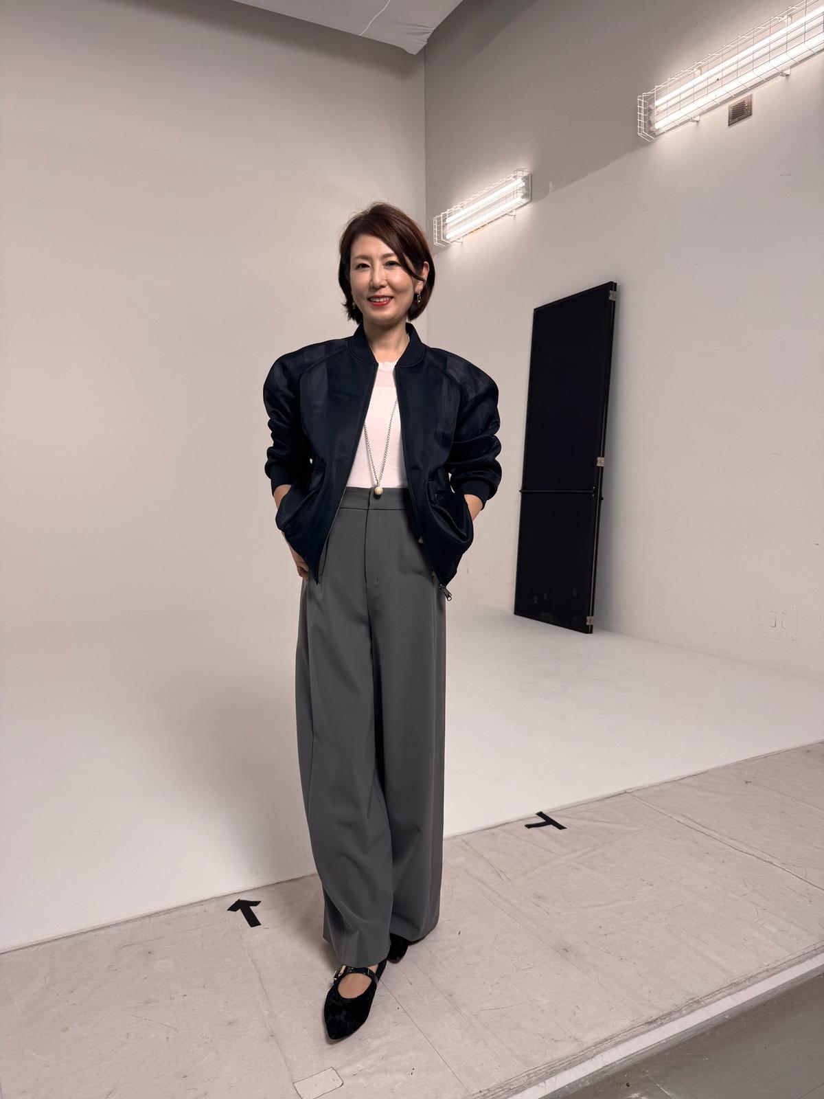 白い壁のスタジオで、グレーのワイドパンツとピンクのトップスに、黒いボンバージャケットを着た女性が立っている。黒いパンプスを履き、右手をポケットに入れている。背景には白い壁と蛍光灯、黒いパネルがある。