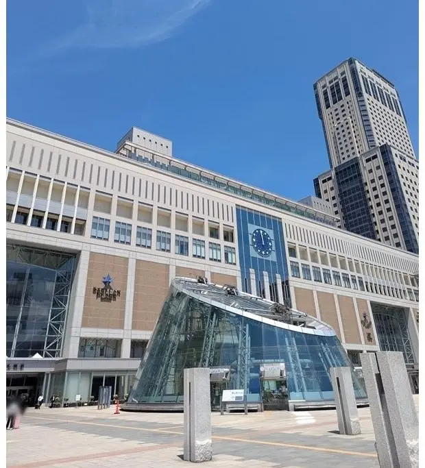 札幌駅周辺の風景。大型商業施設と高層ビルが並び、青空に白い雲が浮かんでいる。建物の外観は白とベージュを基調とし、ガラス張りの構造が特徴的。