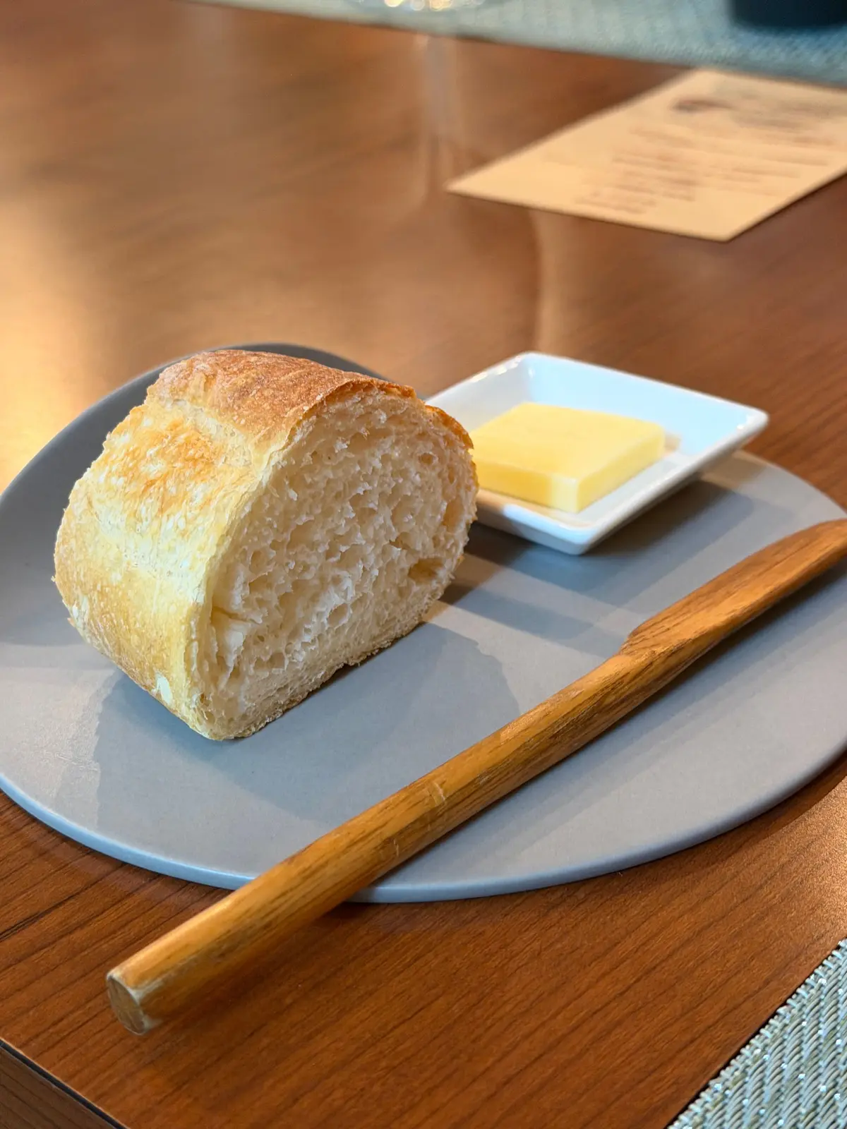 灰色の皿に、焼いたパンの切れとバター、それに木製のバターナイフが置かれている。パンは内側がふっくらとしており、バターは四角い白い容器に入っている。