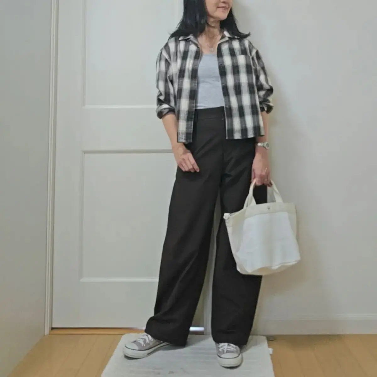 黒いワイドパンツに白いタンクトップ、白黒のチェック柄シャツを羽織った女性。白いトートバッグを持ち、グレーのスニーカーを履いている。背景は白いドア。
