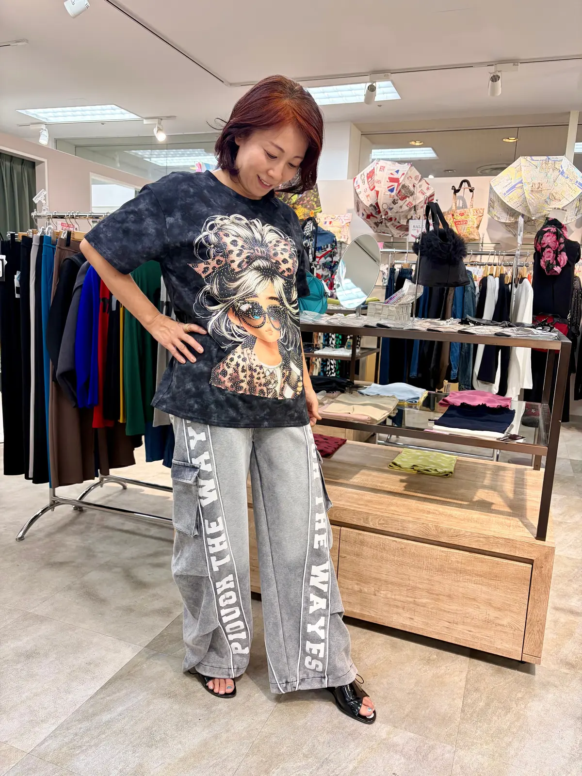 女性が、プリントTシャツと「DO YOUR THING」と書かれたスウェットパンツを着用し、お店で身につけている様子を覗き込んでいる。背景には、傘や衣料品が並んだ棚が見える。