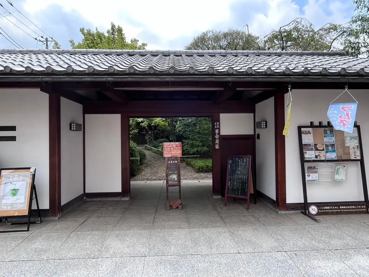 瓦屋根の門から見える庭園。茶色の柱と黒い扉で構成された門の奥には、石畳の通路と緑豊かな植栽が見える。右側には案内板やポスターが設置されている。
