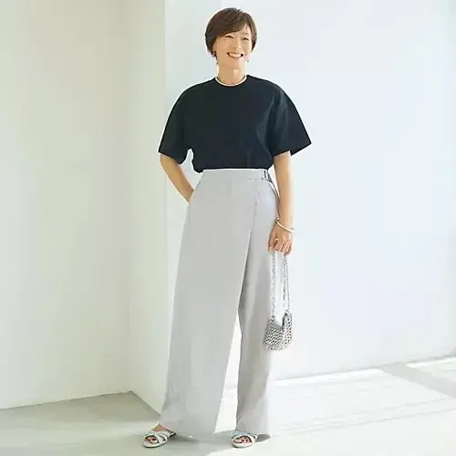 黒の半袖Tシャツとグレーのワイドパンツを着た女性が、白い壁の前で微笑んでいる。右手に小さな編みかごバッグを持ち、足元には白いサンダルを履いている。