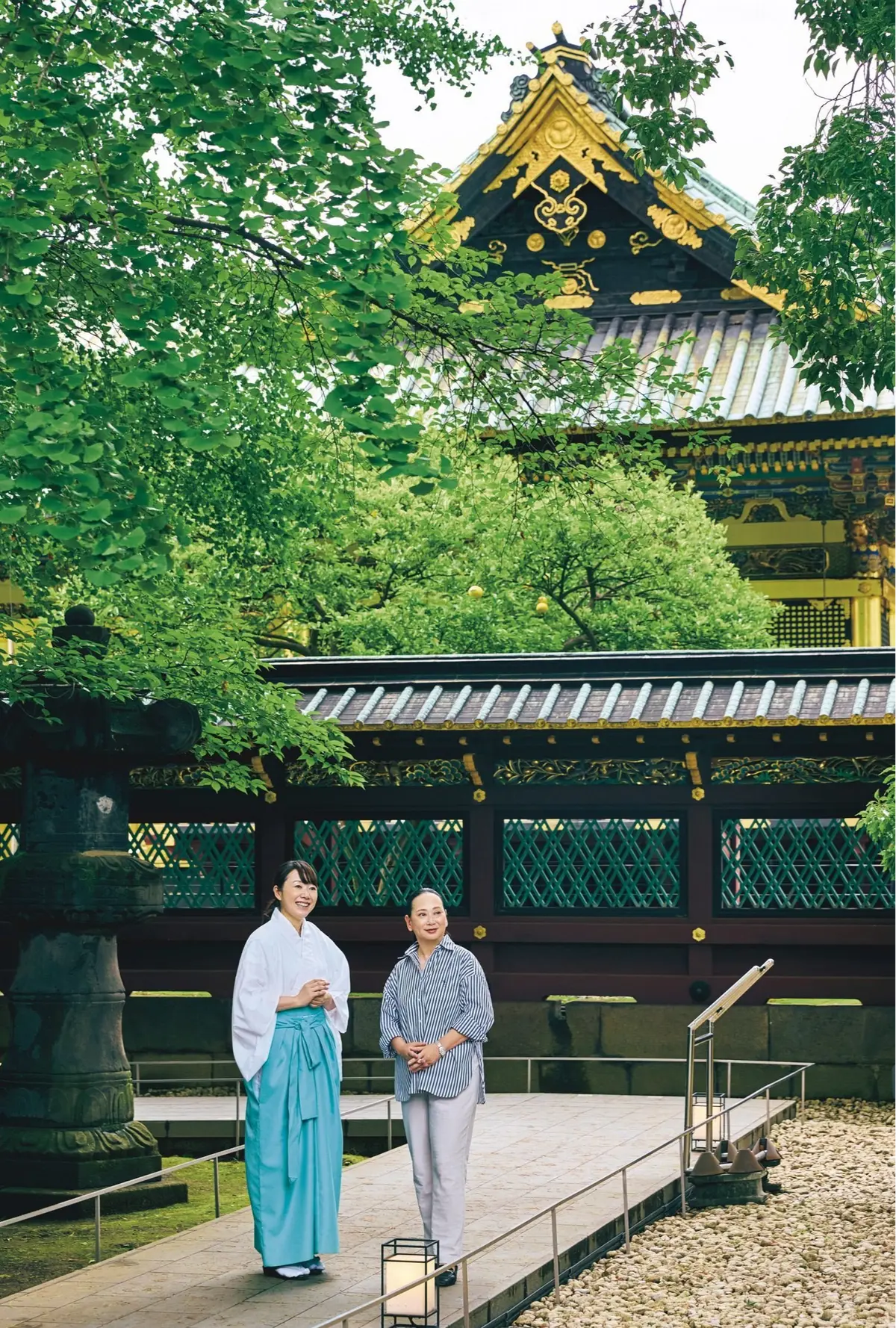 緑豊かな木々と伝統的な日本の神殿を背景に、和装の女性とカジュアルな服装の女性が立っている。手前にランタンが置かれ、通路には手すりが設置されている。