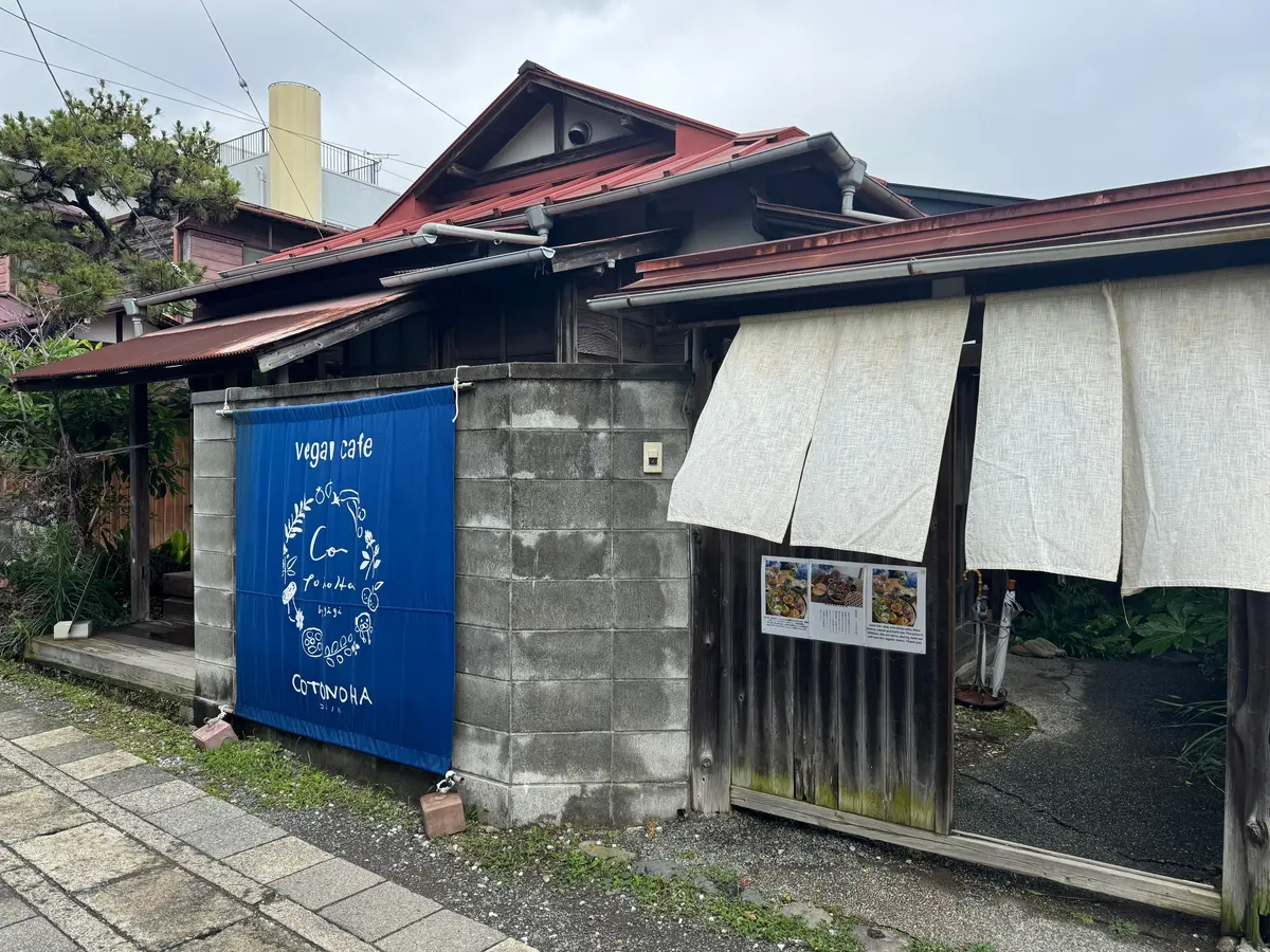 ベジカフェ「コトノハ」の外観。青い看板に店名とロゴ、木製のドア、ベージュのカーテン、メニュー表が設置されている。赤い屋根とコンクリート壁の建物。