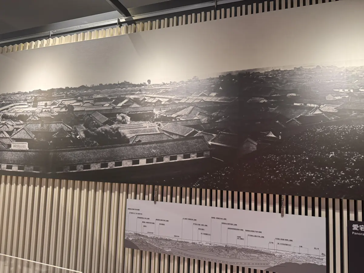 博物館の展示室。黒い壁に、古い街並みの白黒写真が広範囲に展示されている。写真の下には、地形図のようなイラストと、日本語の解説文が配置されている。右下には、白い木製の仕切りが立っている。