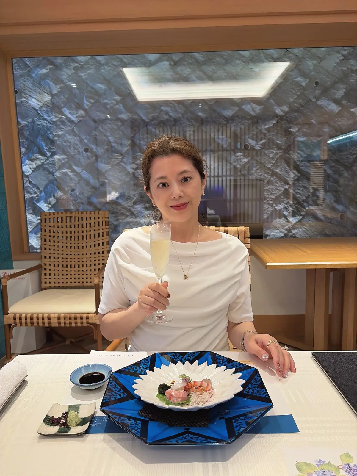 女性が白い服で、シャンパングラスを手に持つ。前に青い花模様のプレートに刺身とワサビが並び、小さなボウルにソースが置かれている。背景には石の壁が映る。