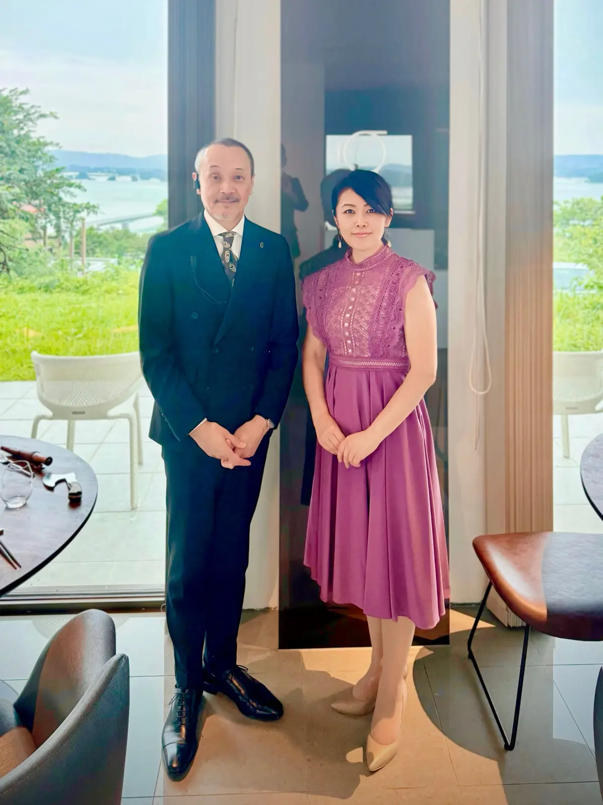 男性と女性が室内にて並んで立つ。男性は紺色のスーツ、女性は紫のドレスを着用。背景には窓から見える景色と椅子がある。