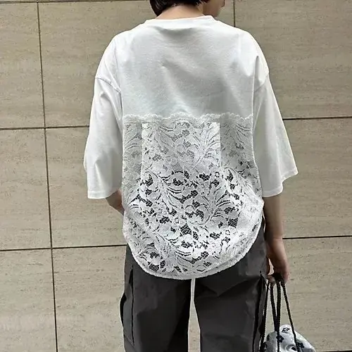 白のオーバーサイズTシャツ。後ろ身頃下部に白いレースがあしらわれている。男性が着用し、黒いパンツとトートバッグを持っている。背景はベージュのタイル壁。