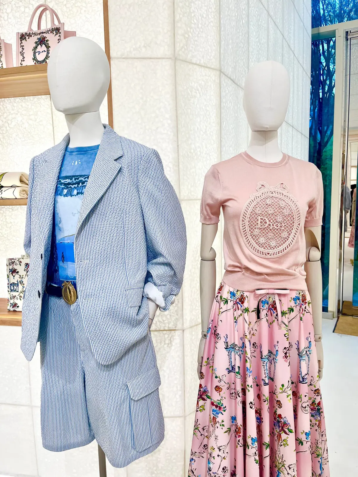 ディオールのディスプレイ。左には青いストライプのジャケットとショートパンツのセットアップ、右にはピンクのTシャツと花柄のフレアスカート。背景にはディオールのバッグや装飾品が飾られている。