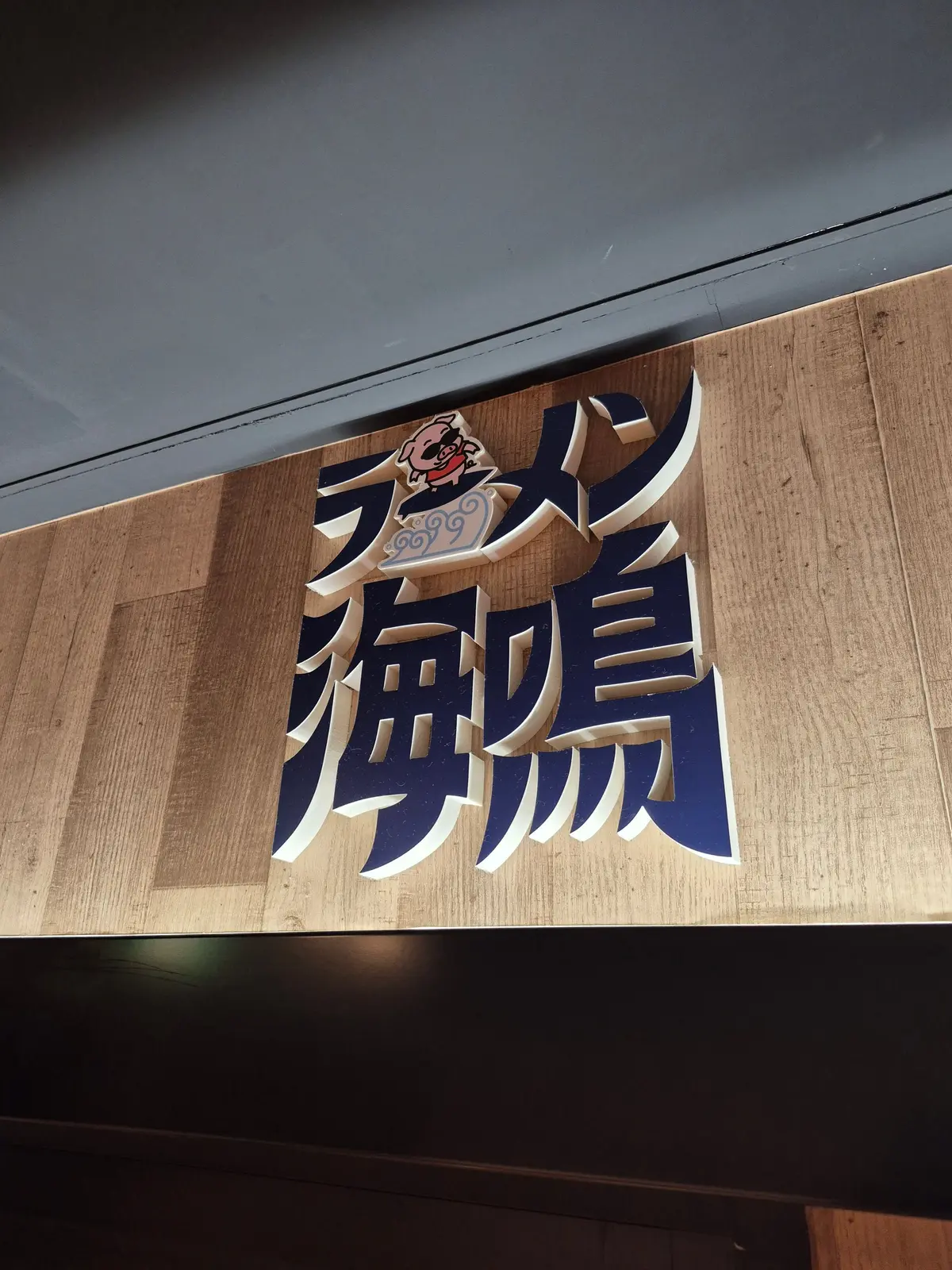 木製の背景に、青地に白字で「豚願」と書かれた看板。上部には豚のイラストが描かれている。