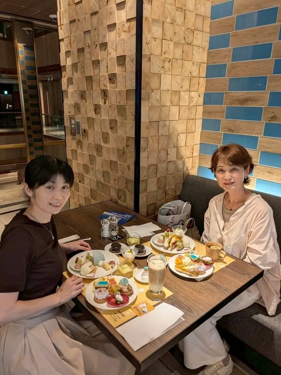 木製のテーブルに多彩なデザートと飲み物が置かれたカフェで、2人の女性が笑顔で座っている