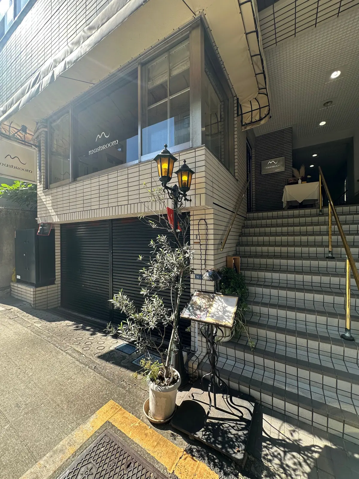 灰色の建物に面した、階段と植栽のある入り口。看板には「mashroom」の文字。手すりの横にはメニュー立てが置かれている。