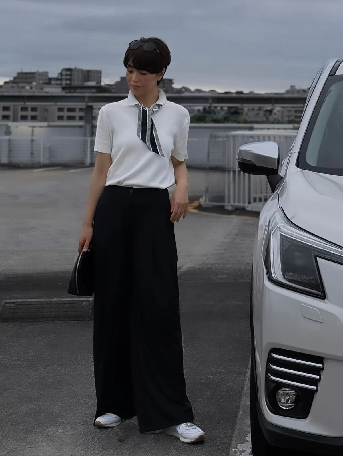 女性が白い半袖シャツに黒のスカート、黒のネクタイを着用し、頭にサングラスをかけ、右手に黒のクラッチバッグを持ち、白いスニーカーを履いて駐車場に立っている。右側に白い車が停まっている。