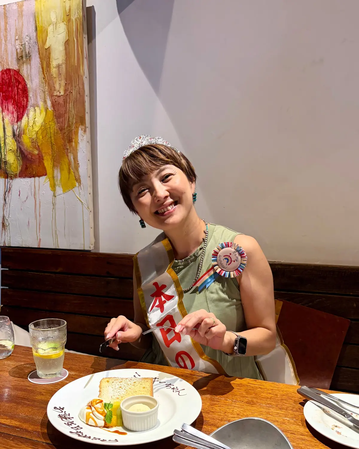 笑顔の女性が、赤い絞り飾りを首にかけ、テーブルのケーキを見ている。茶色の短髪で、頭には小さなティアラを付けている。背景には抽象画が飾られ、テーブルには飲み物と食器がある。