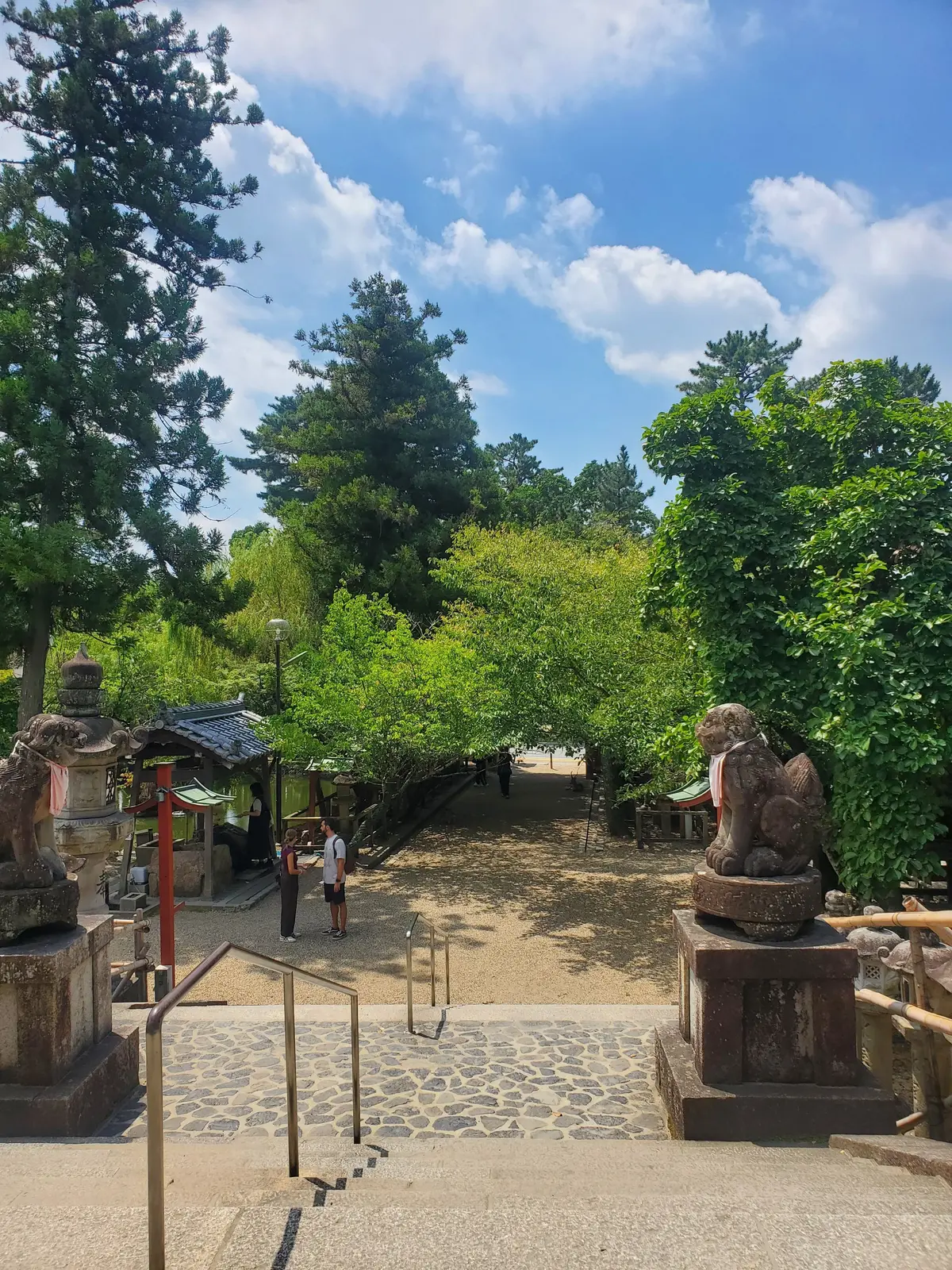 石像を配した階段状の参道。緑豊かな木々が両側に茂り、上部には青空と雲が見える。中央には人が数名歩いている。