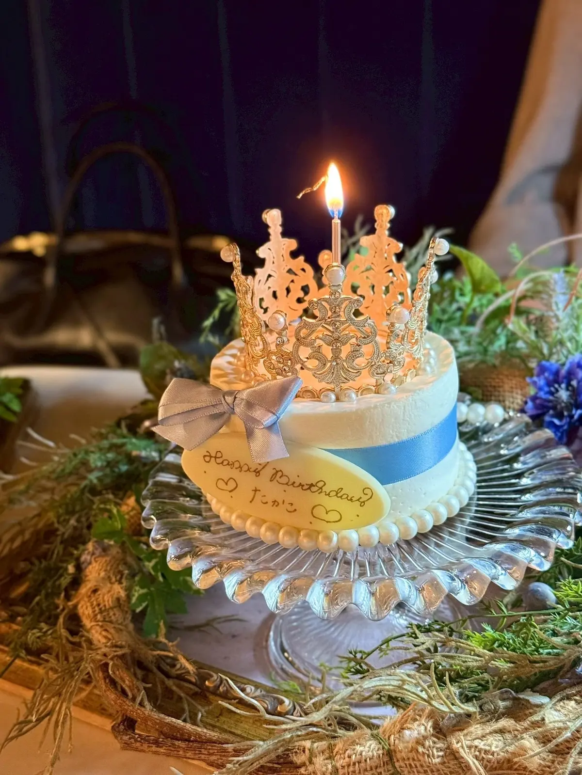 梅田ブルーバードの誕生日サプライズで登場した華やかなティアラケーキ　ベージュと青のツートーンのケーキに、精巧な金色の王冠が乗せられ、一本のキャンドルが点灯している。ケーキの上には「ハンナ・ウィンドロウ・テイラー」と書かれたエレガントなプレートがあり、シルバーのリボンが飾られている。