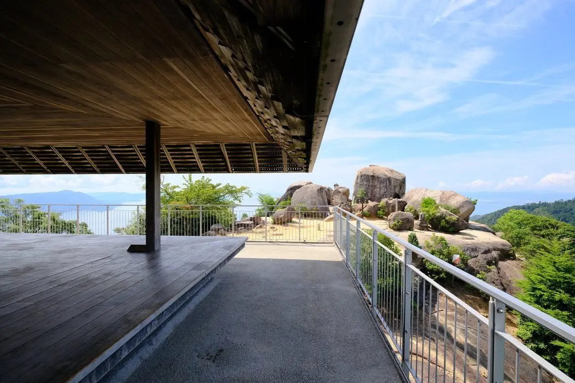木製の屋根が広がる展望台から、緑豊かな丘陵地帯と海を見渡せる風景。手すりが設置された通路が岩場を通り、遠景には島々が連なる。青空に白い雲が浮かぶ。
