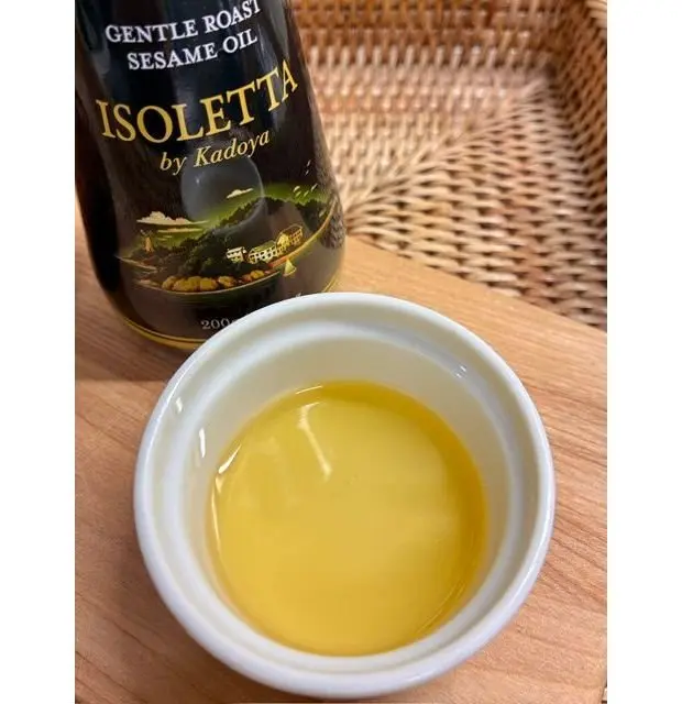木製のテーブルに置かれた、カドヤの「イソレッタ」と書かれたローストゴマ油の瓶。瓶の横には白い器に油が注がれており、光を受けて淡い黄色く輝いている。