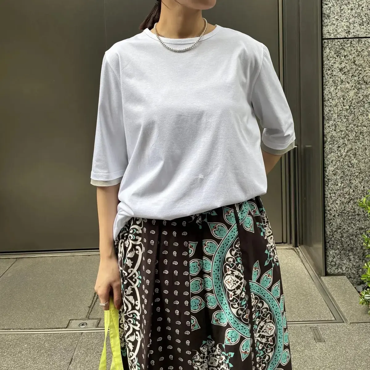 白の半袖トップスと緑と黒のペイズリー柄スカートを着た女性が、建物の前で立ち、左手には黄色いカバンを持っている。