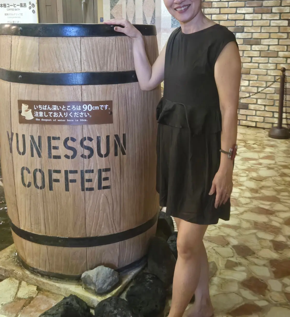 木製の巨大な樽の横に立つ女性。樽には「Yunessun Coffee」の文字が大きく書かれている。女性はグレーのワンピースを着ており、右手を樽に添えている。背景にはレンガの壁と木製の看板が見える。