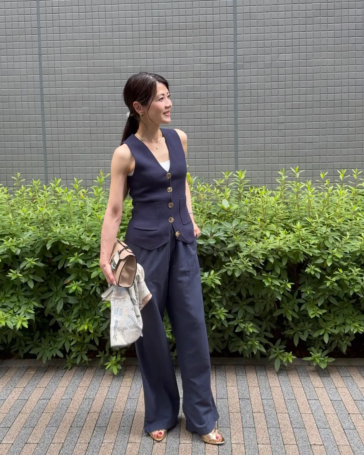 女性が紺色のベストとワイドパンツのセットアップを着て、屋外で微笑んでいます。彼女は左手に小銭入れと布製の袋を持っています。背景には緑豊かな低木があり、グレーのタイル壁が並んでいます。
