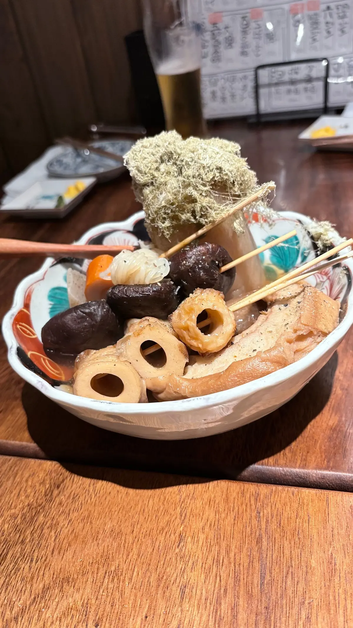 串に刺さった様々な種類の煮物。レンコン、こんにゃく、椎茸、かまぼこなどが盛り付けられ、乾燥海苔が添えられている。白い器に盛り付けられ、木製のテーブルに置かれている。