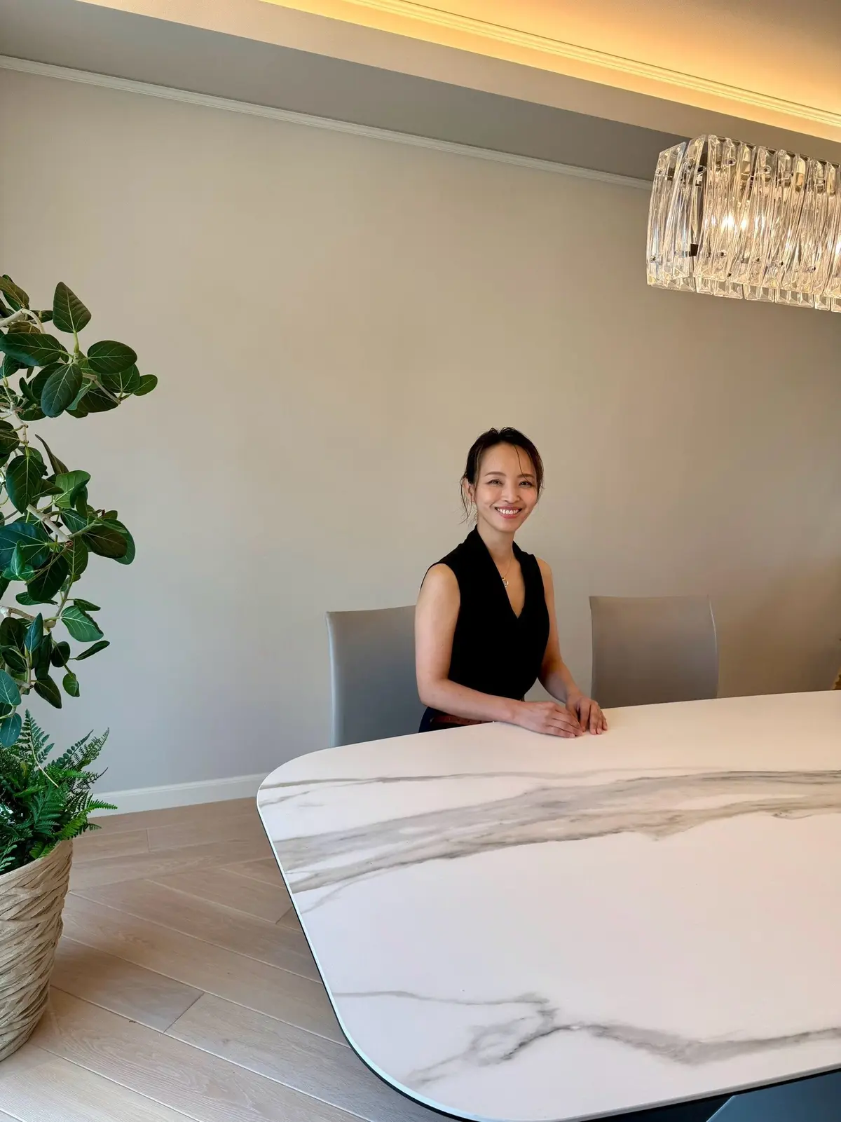 明るい部屋で、白い大理石のテーブルに座る女性。黒いタンクトップを着て、笑顔でこちらを見ている。左には緑の植物が置かれ、天井にはシャンデリアがある。