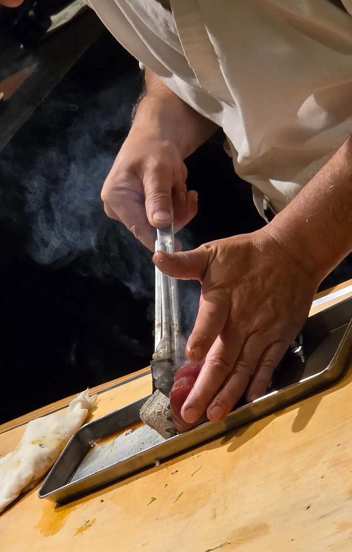 寿司職人が、赤いマグロを薄く削る様子。木製のカウンターに金属製のトレーがあり、削り出したマグロが乗っている。背景には、他の食材や調理器具が見える。