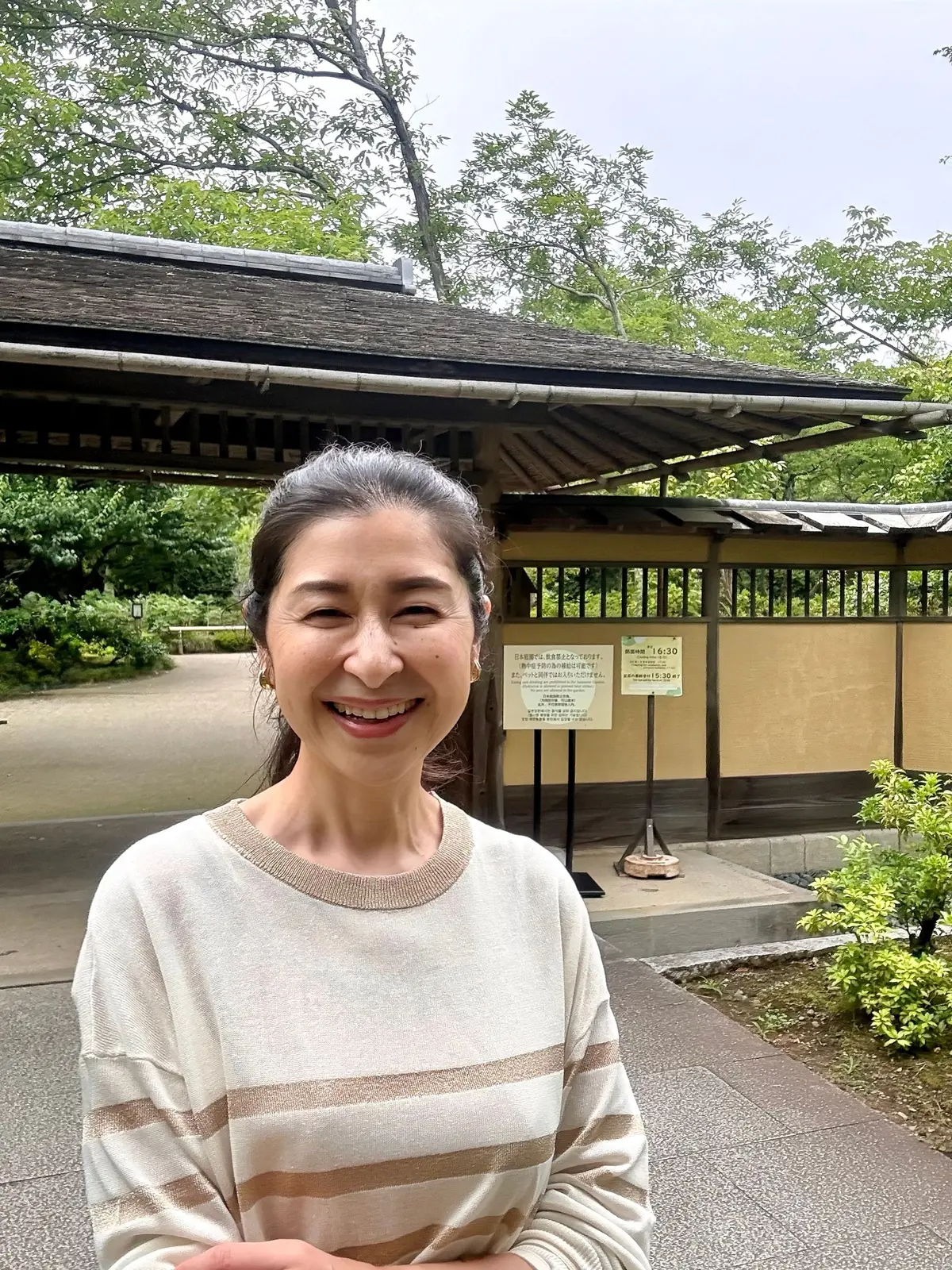 国営昭和記念公園、日本庭園、ベージュと茶色の縞模様のセーターを着た女性が、木造の建物と緑の木々が背景にある庭園で微笑んでいる