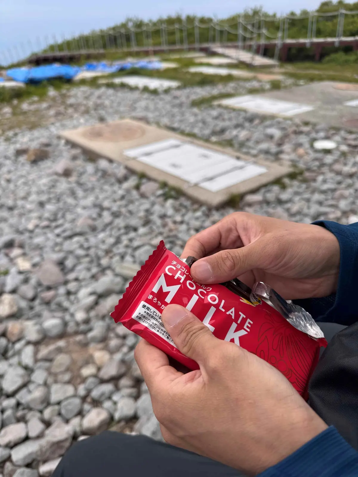 手は赤いチョコレートミルクバーを部分的に開封し、岩の多い地面の上に置かれた木製の台座と、遠くに緑の芝生とフェンスが見える背景が広がっています。