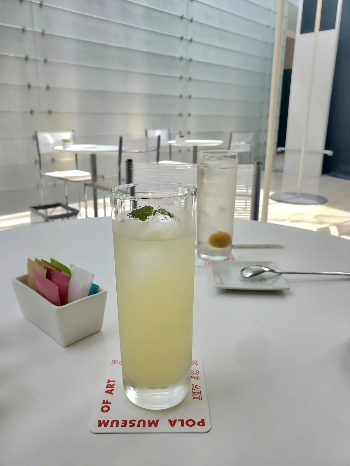 白いテーブルに置かれた、レモン風味のドリンクと水、ナプキン、POLA MUSEUMのロゴが入ったコースター。背景には白い壁と窓が見える。