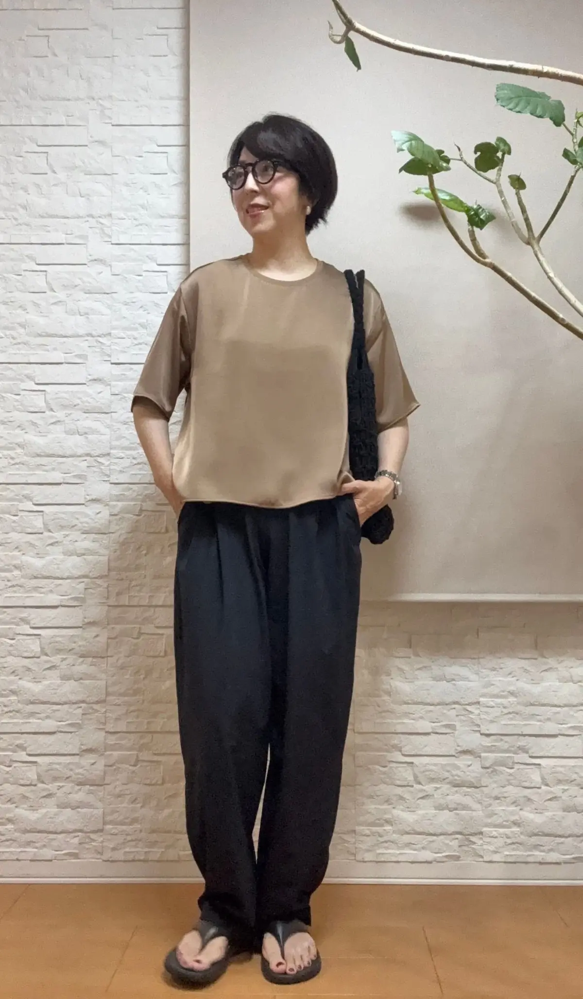 ベージュの半袖シャツと黒のワイドパンツを着た女性が、壁に寄りかかり、右手に黒の鞄を持っている。白いレンガの壁と緑の植物が背景にある。