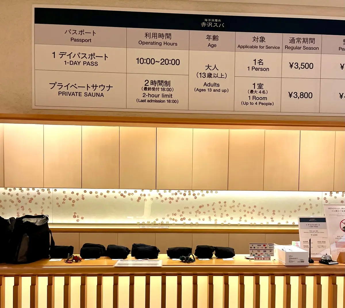木目調の待合スペース。黒板に料金表が掲示され、黒い椅子が並んでいる。奥には白い壁に金色の模様が施されている。