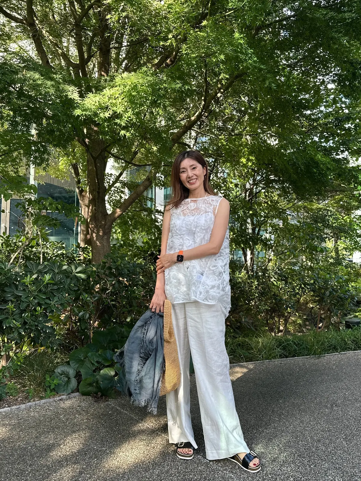 白いワイドパンツとレースのトップスを着た女性が、緑豊かな公園で微笑んでいる。左腕にデニムのジャケット、右腕にバッグを持ち、黒いサンダルを履いている。背景には現代的な建物が見える。