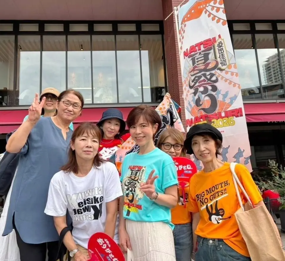 笑顔のグループ写真。6人の女性が並んでおり、それぞれ異なる色のTシャツを着ている。背景には、窓や建物、そして「Natsu Matsuri 2026」と書かれた旗が飾られている。