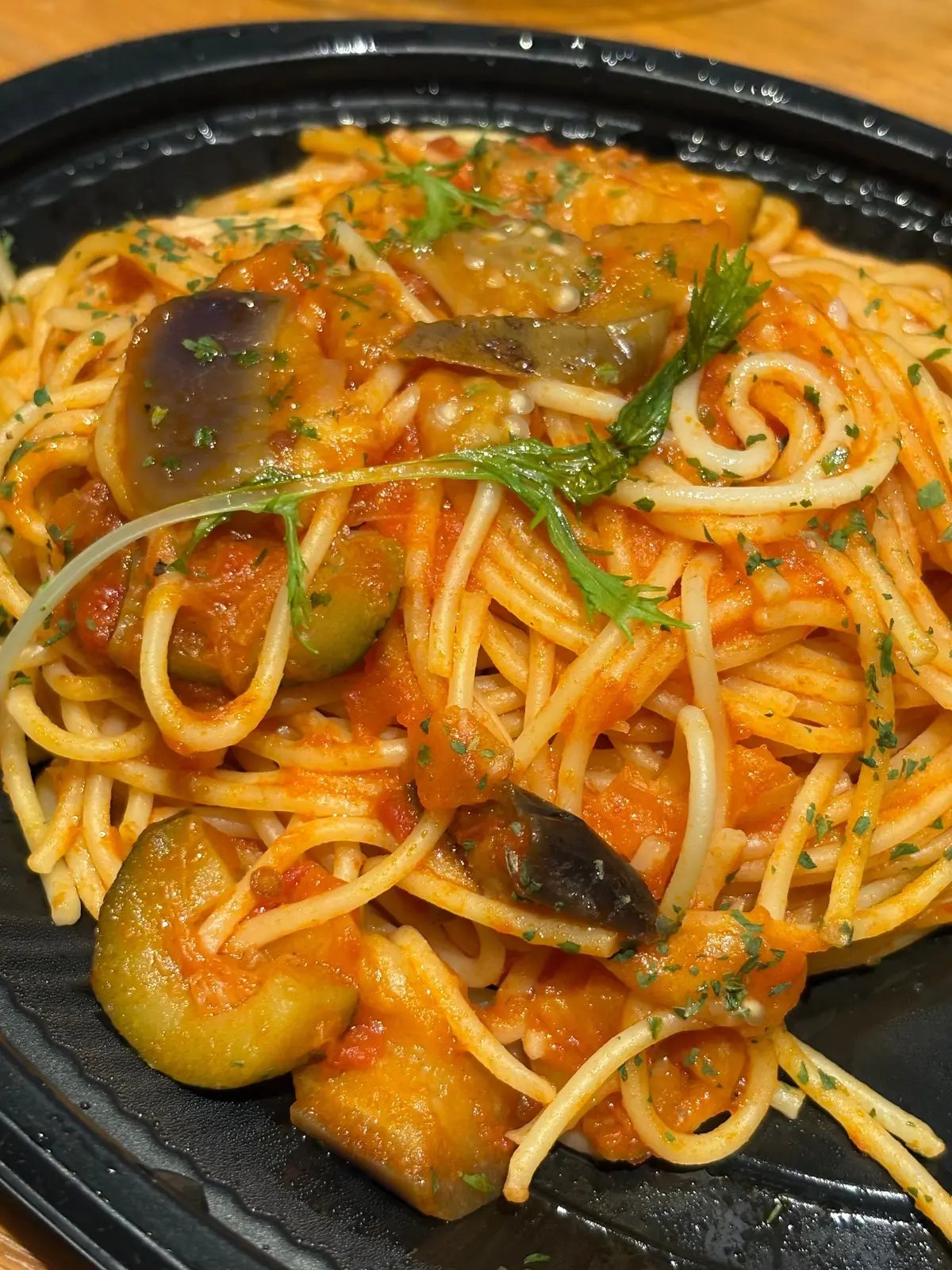 黒い皿に盛り付けられた、トマトソースのスパゲッティ。揚げたナスやオリーブ、緑のハーブが添えられている。