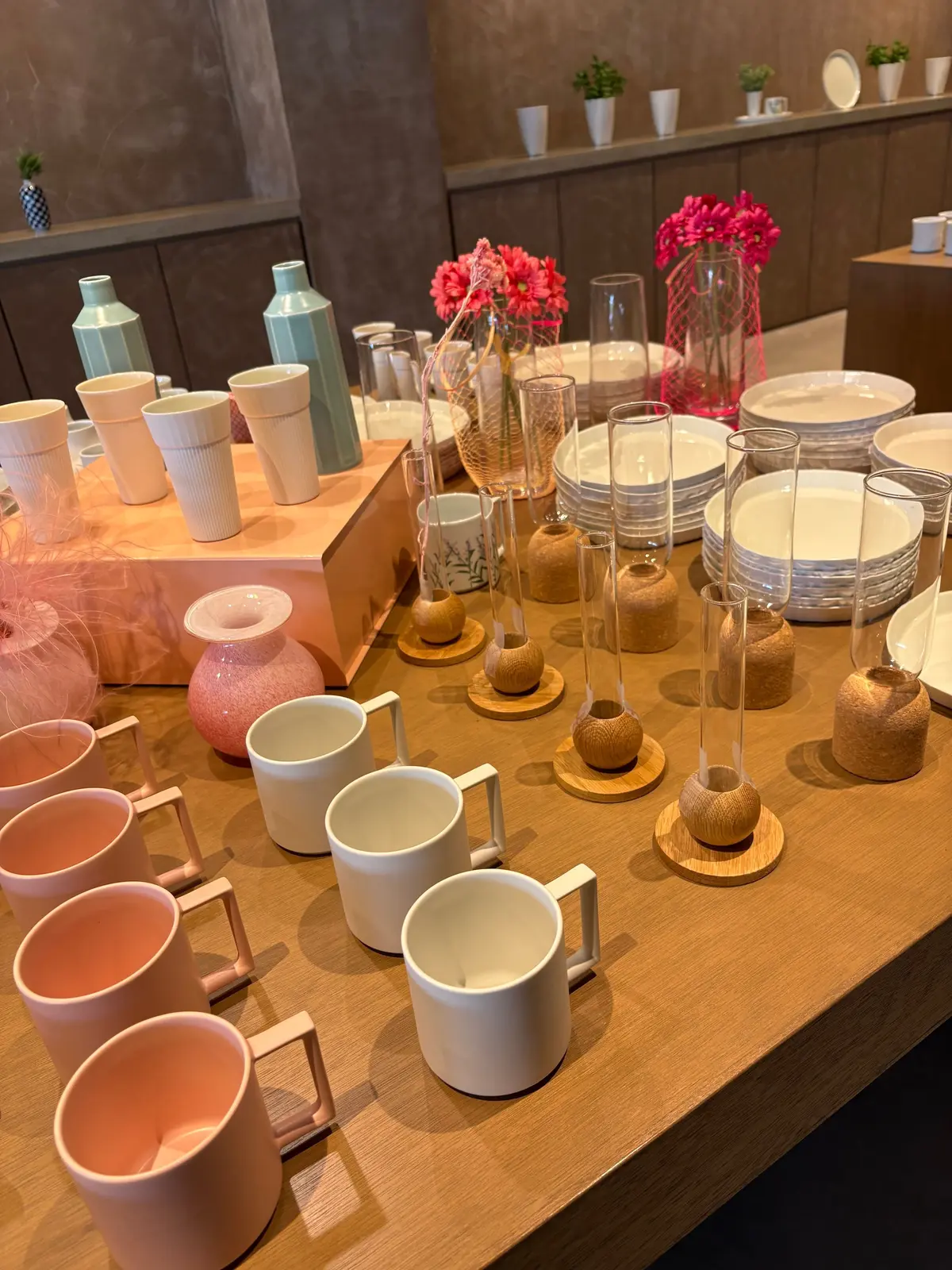 木製のテーブルにピンクと白の陶器マグカップ、ガラスカップ、花瓶が並び、背景に小花の入った花瓶と棚に置かれた小物も見える