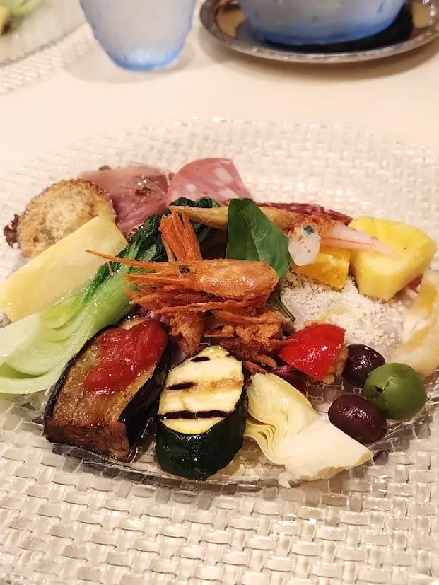 ガラス皿に盛り付けられた、彩り豊かな前菜の盛り合わせ。グリル野菜、魚介、チーズ、オリーブ、ハーブなどがバランスよく配置されている。