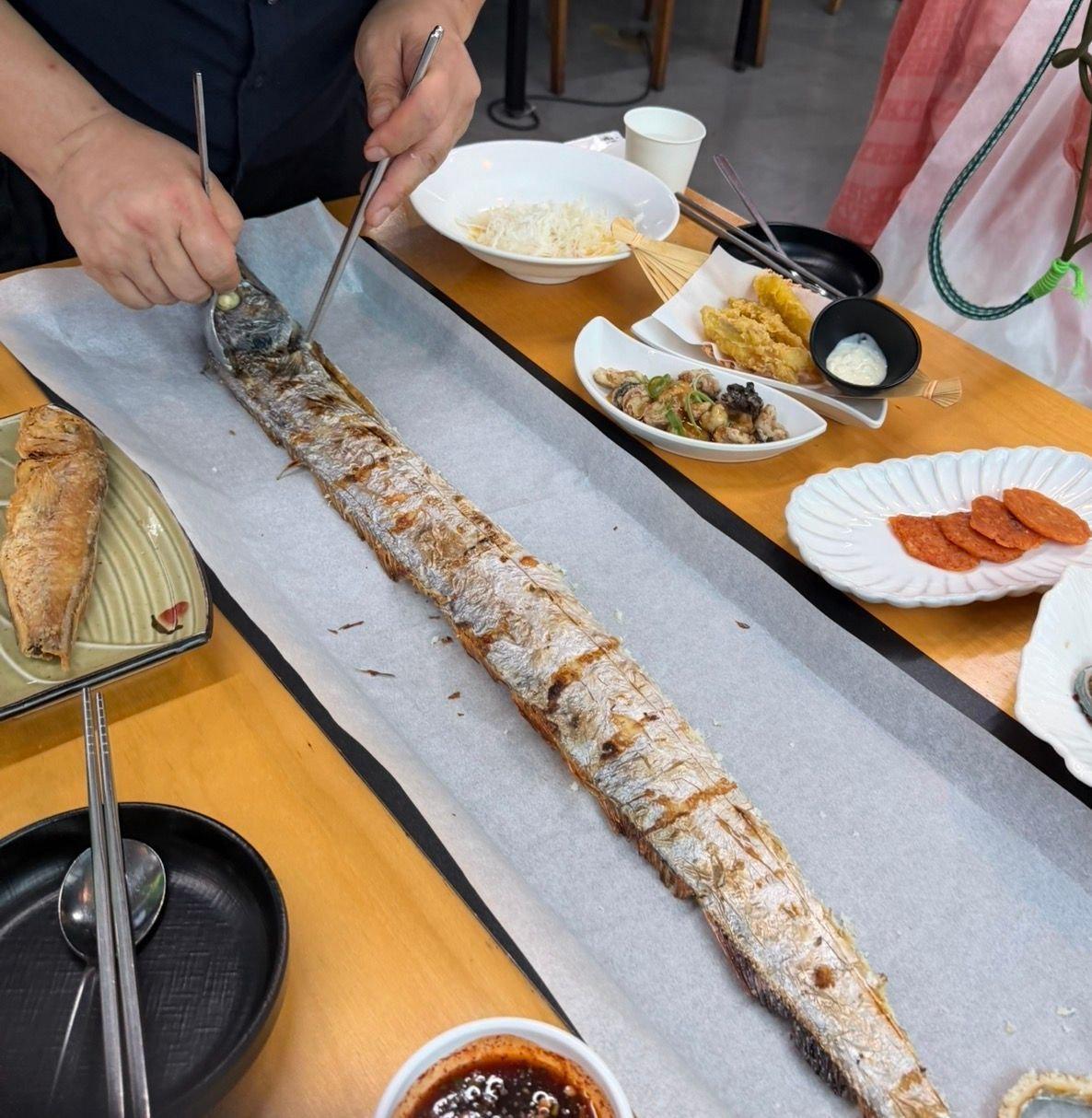 テーブルに置かれた長細い焼き魚。人の手で取り分けられており、キャベツの千切り、天ぷら、ソーセージなどの料理も見える。