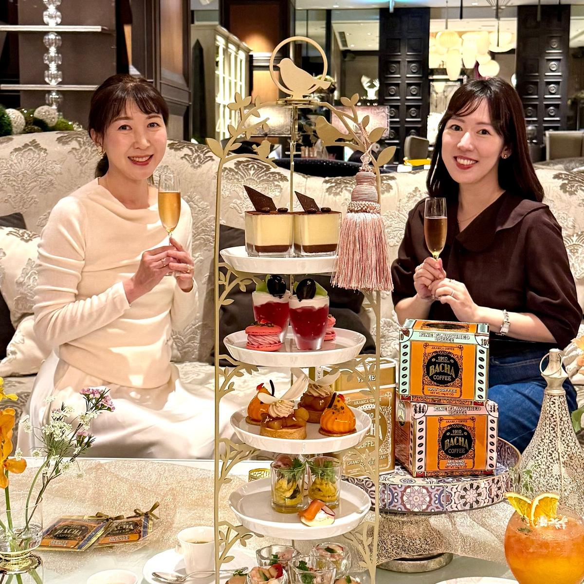 バッハコーヒーのカフェでの写真。2人の女性がシャンパングラスを持ち、豪華なスイーツの3段ケーキを囲んでいる。背景には金色の装飾が施された内装が見える。