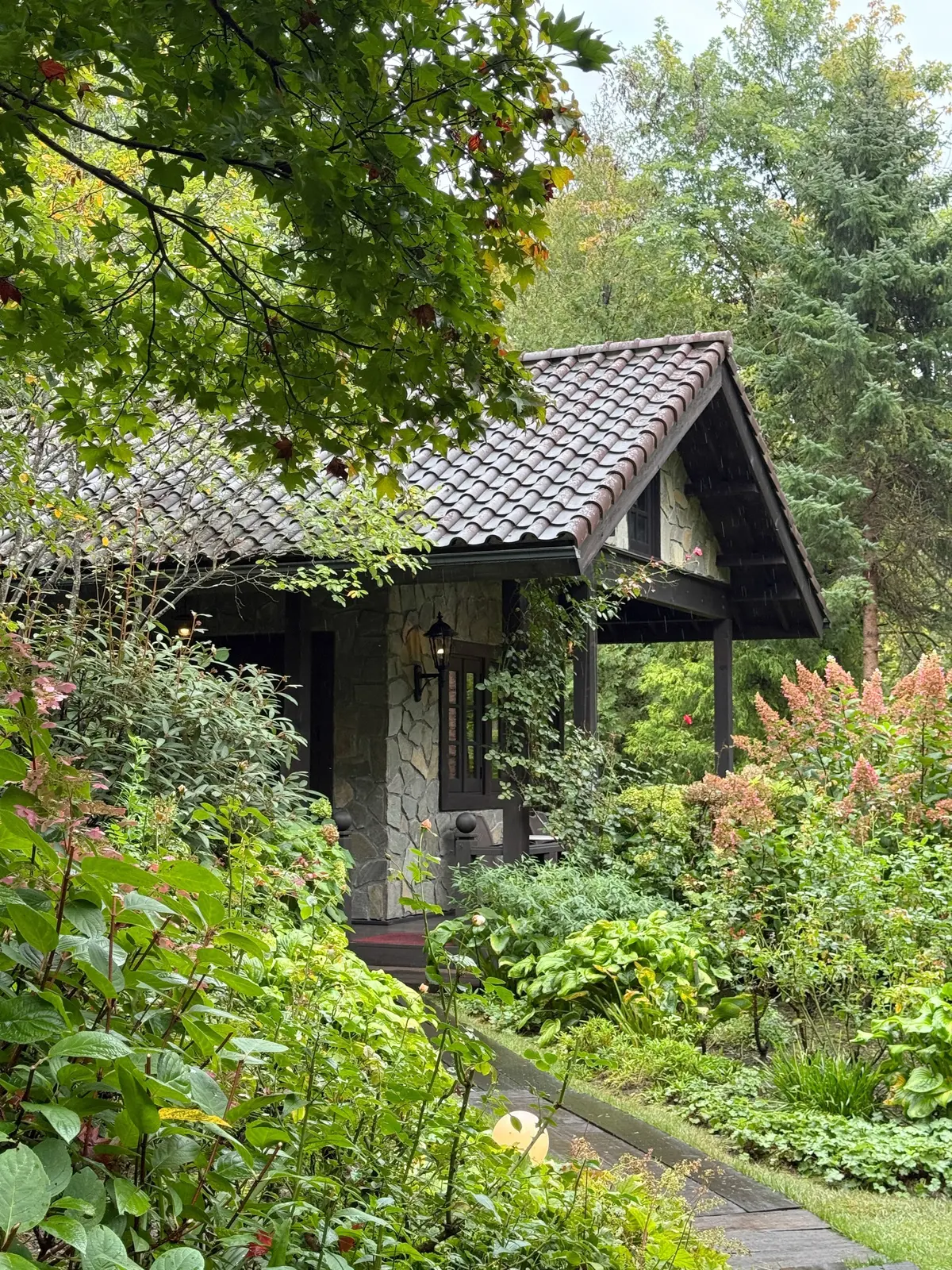 緑豊かな植物に囲まれた、石造りの可愛らしい小屋。屋根は赤茶色の瓦で、入り口にはランタンが灯っている。木製の庇が設けられ、蔦が絡まっている。