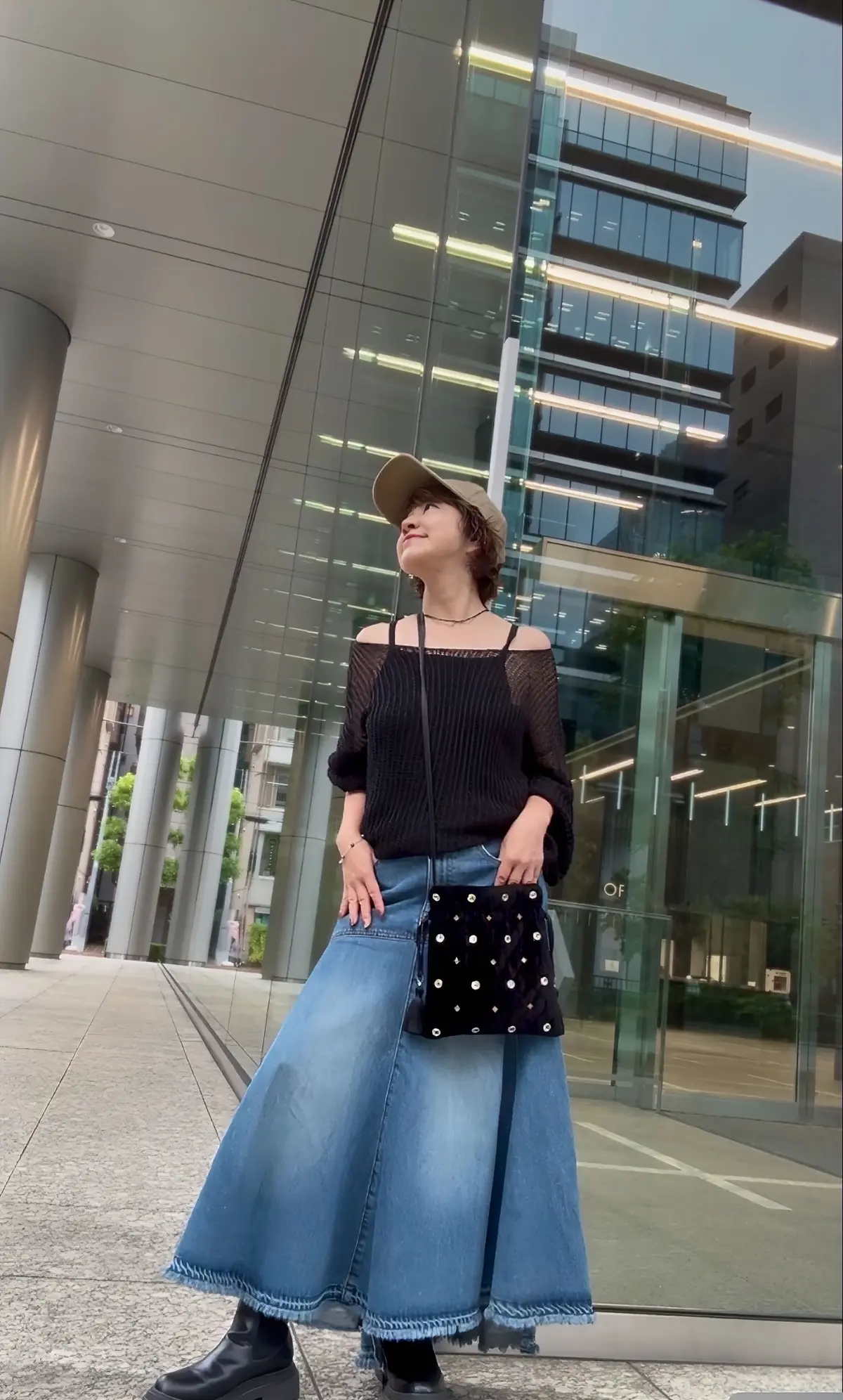 黒いニットトップスとデニムスカートを着用した女性が、都会のビルの前でポーズをとっている。キャップをかぶり、黒いブーツを履いている。手には小さな黒いバッグを持っている。