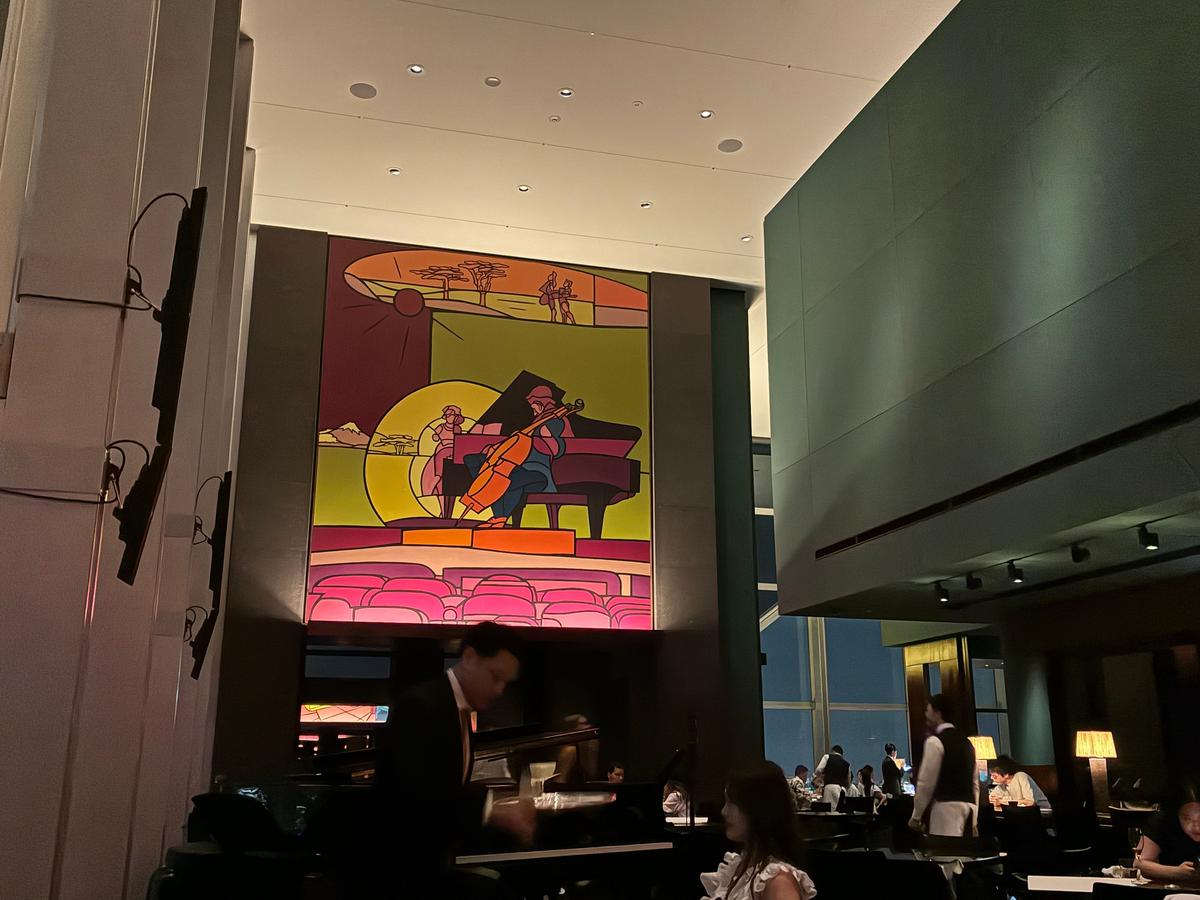 高級レストランの内部。壁にはジャズバンドをモチーフにしたイラストが飾られ、手前にはグランドピアノと数人の客がいる。背景には窓から夜景が見える。