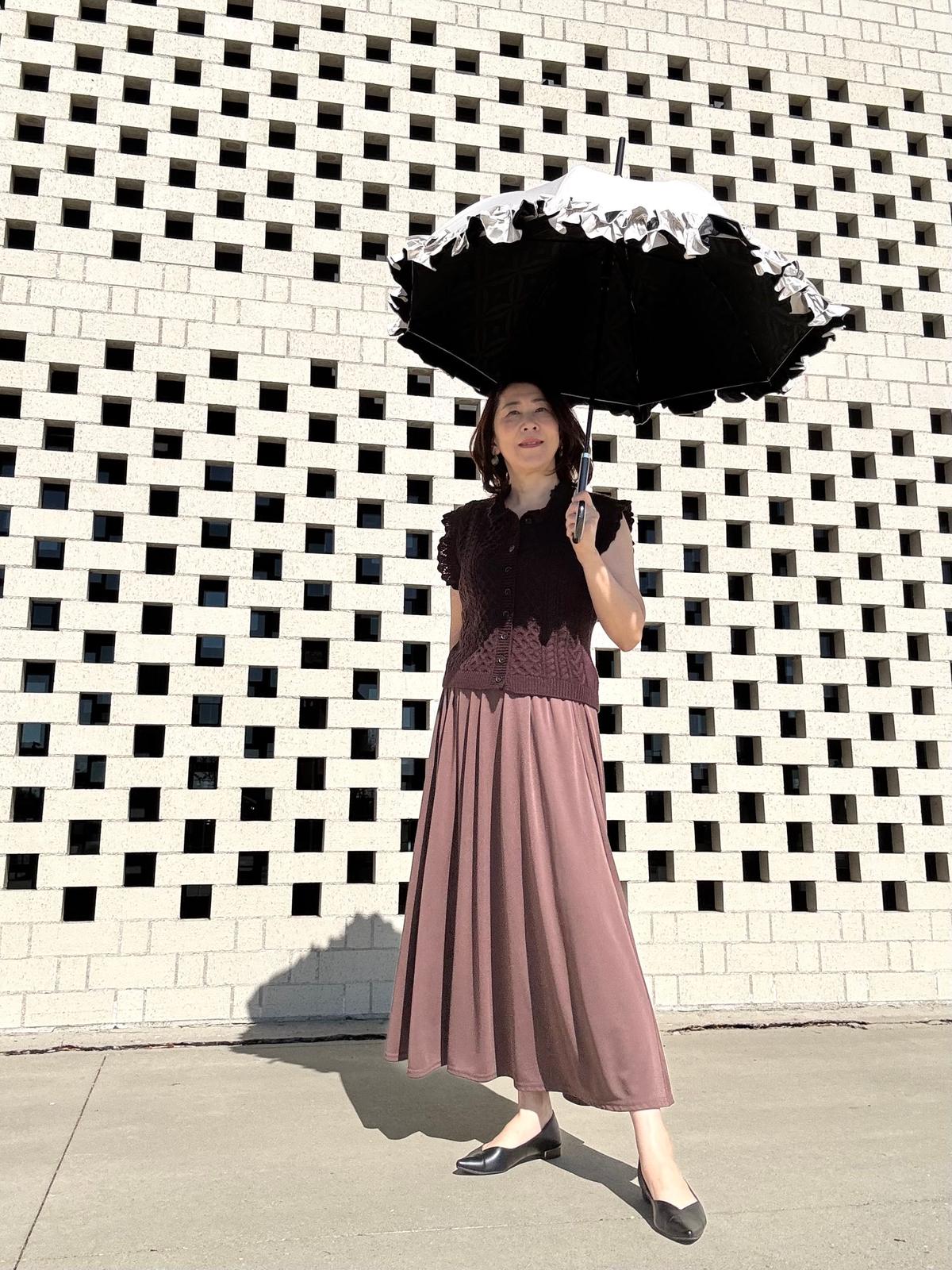 黒い傘をさす女性が、白い幾何学模様の壁の前に立っている全身写真。茶色のトップスとスカートを着用し、黒いパンプスを履いている。
